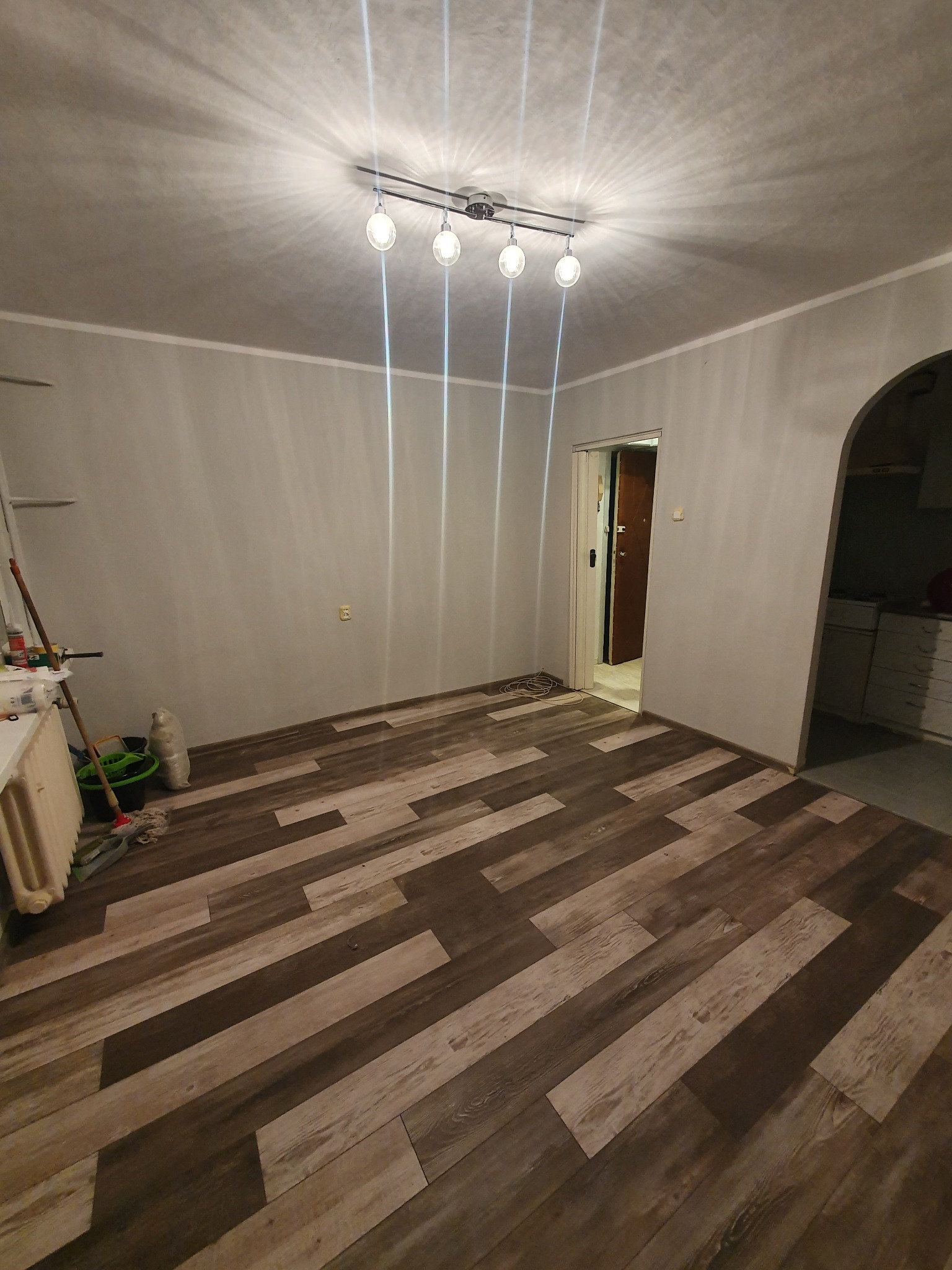 Wnętrze pokoju po remoncie z panelami podłogowymi imitującymi drewno, widoczny grzejnik, wiadro z mopem i otwarte drzwi do innego pomieszczenia, oświetlone lampą sufitową z kilkoma żarówkami.