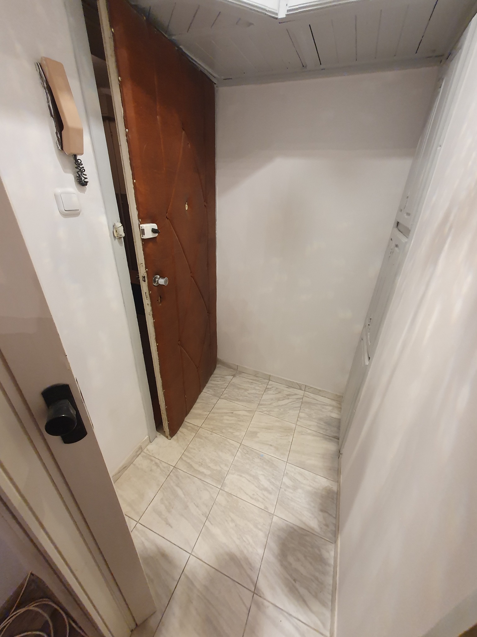 Wnętrze przedpokoju z brązowymi drzwiami wejściowymi, białymi ścianami i jasną, kwadratową podłogą z płytek. Na ścianie widoczny beżowy domofon i włącznik światła.