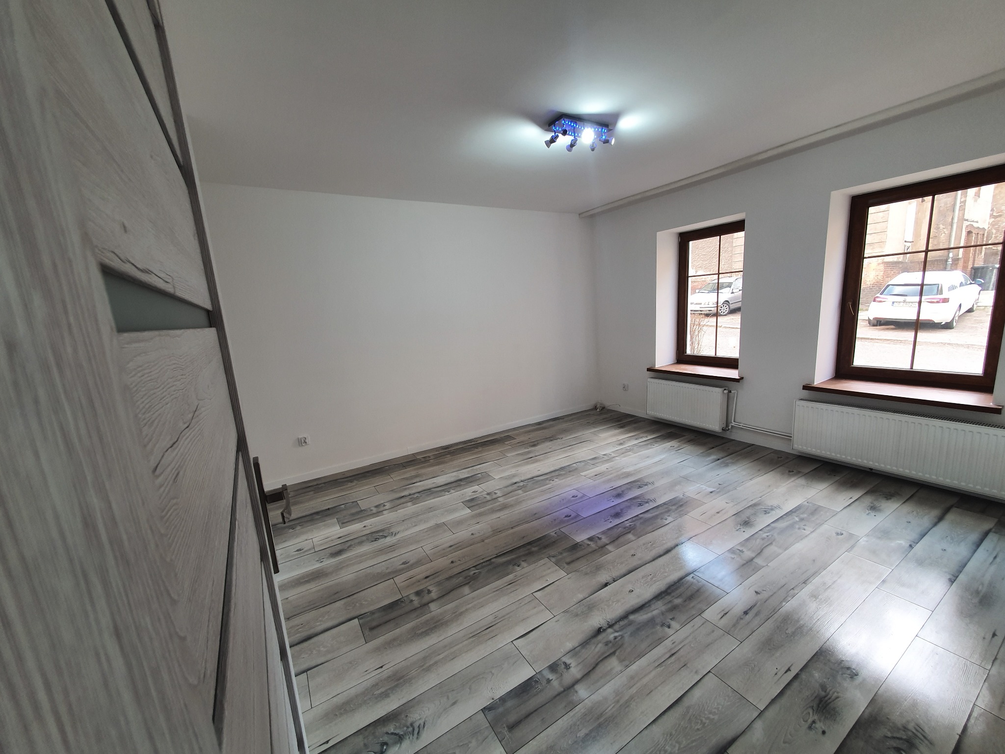 Pusty pokój po remoncie z szarymi panelami podłogowymi imitującymi drewno, białymi ścianami, dwoma oknami z brązowymi ramami i grzejnikami pod nimi, oraz nowoczesną lampą sufitową z niebieskimi...