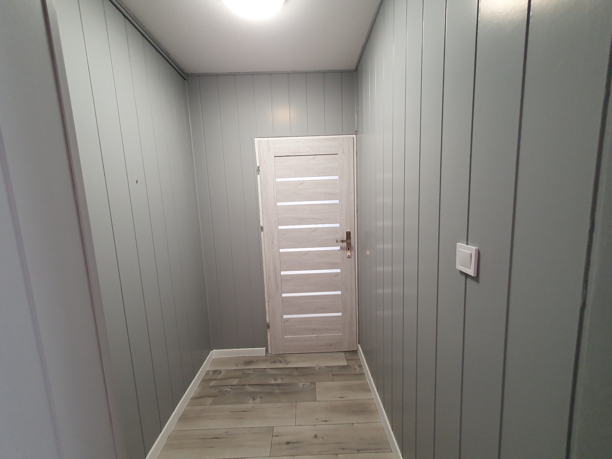 Szare panele ścienne z pionowym wzorem, drewnopodobne płytki podłogowe i białe drzwi z poziomymi przeszkleniami w wąskim korytarzu podświetlonym lampą sufitową.