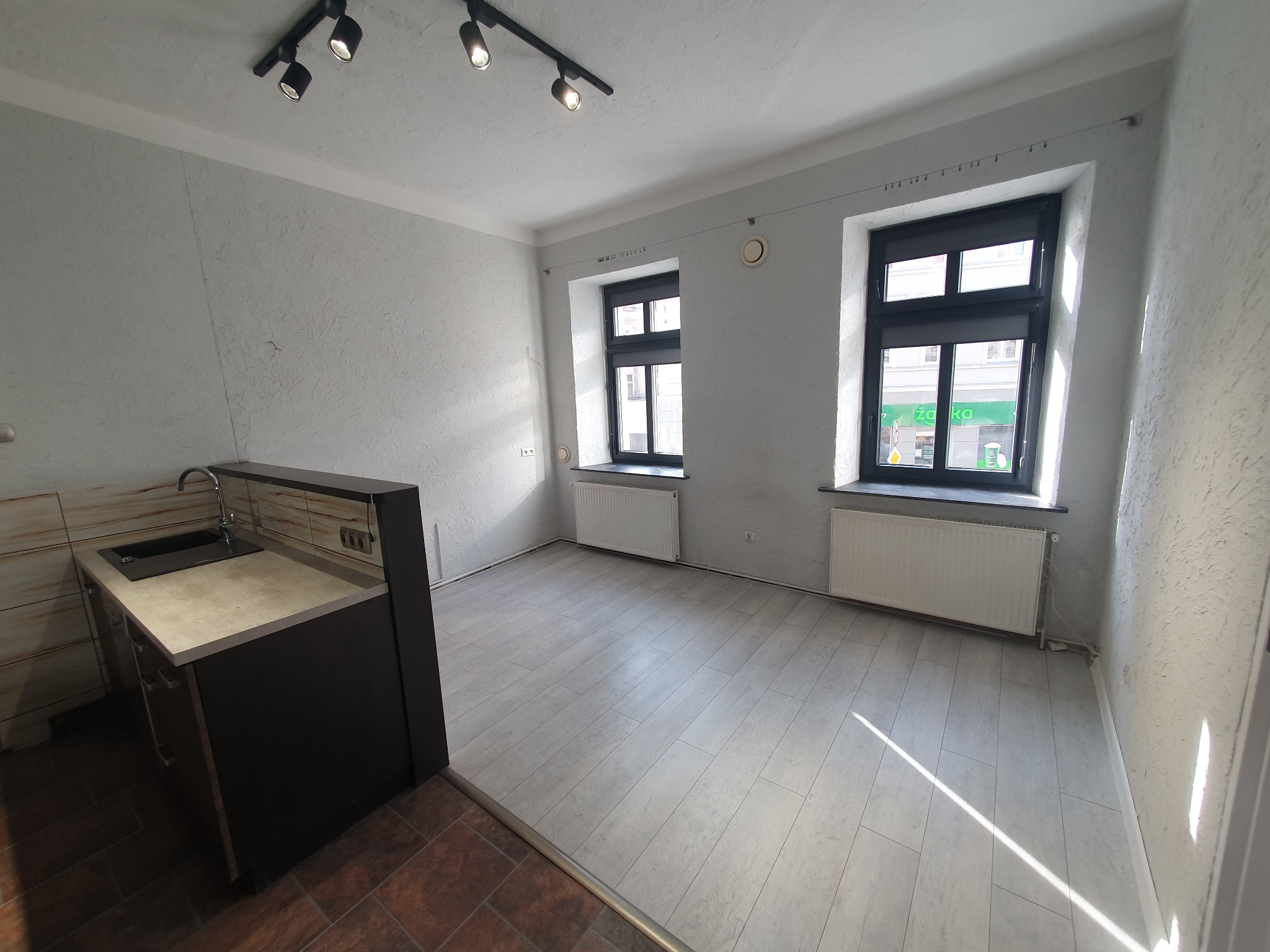 Pusty pokój z dwoma oknami, grzejnikami, panele podłogowe w jasnym odcieniu drewna i fragmentem aneksu kuchennego z ciemnymi szafkami, widoczne lampy sufitowe na szynie.