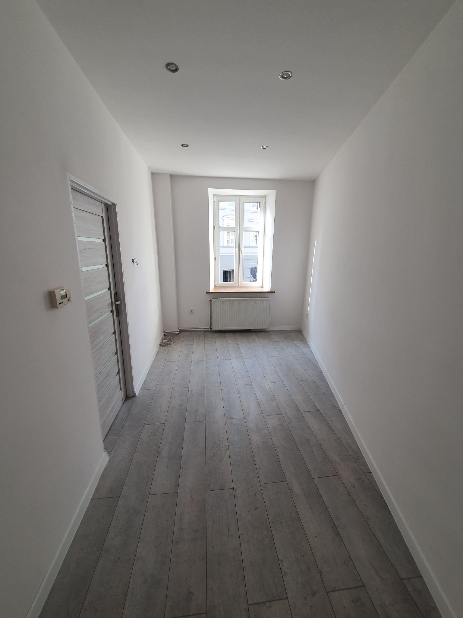 Puste, odnowione pomieszczenie z szarymi panelami podłogowymi, białe ściany, okno z grzejnikiem, szare drzwi z poziomymi wstawkami i widoczne punkty oświetleniowe na suficie.