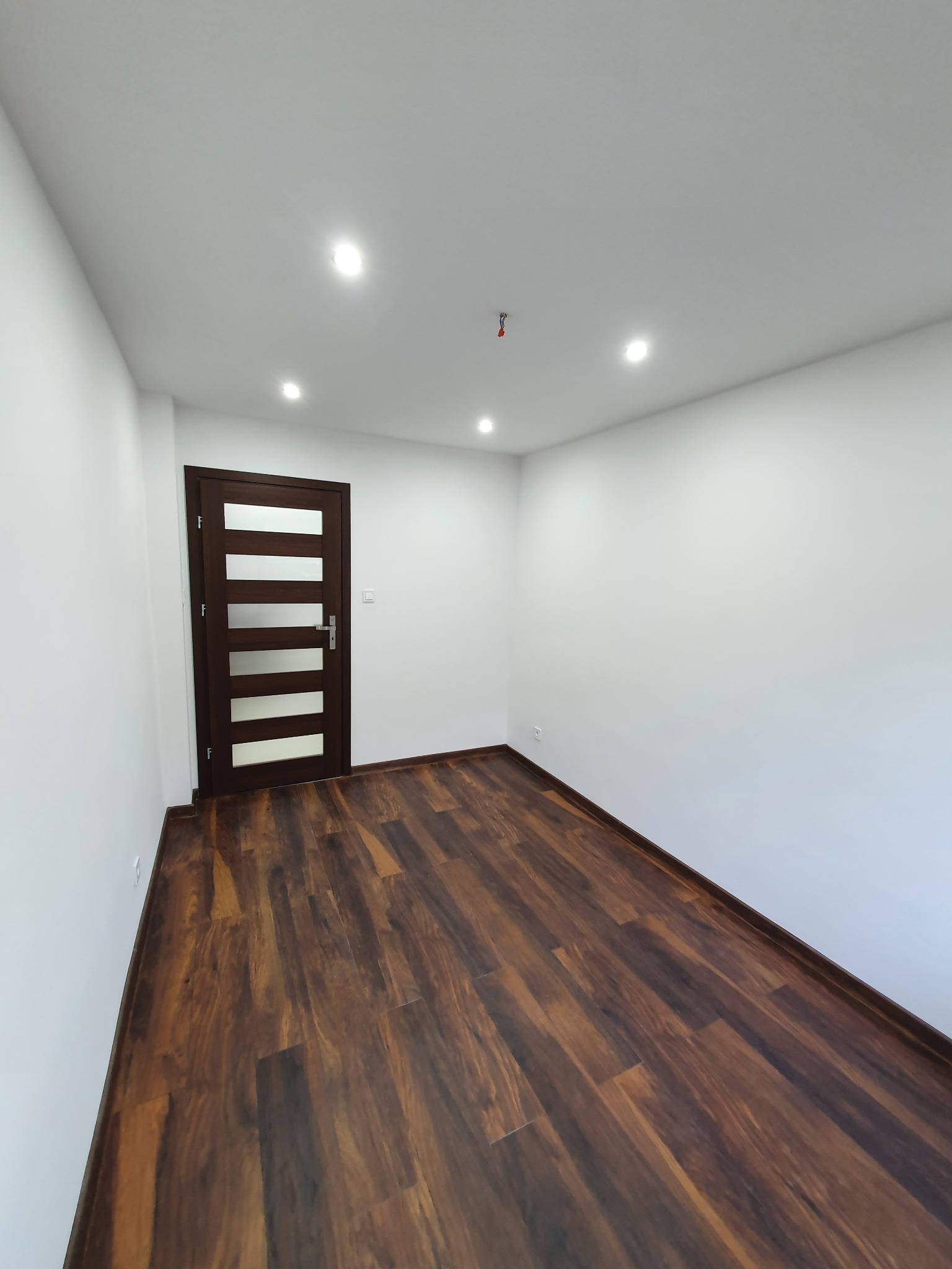 Pusty pokój po remoncie z ciemnobrązową podłogą z paneli, białe ściany i sufit z oświetleniem punktowym oraz ciemne drzwi z przeszkleniami.