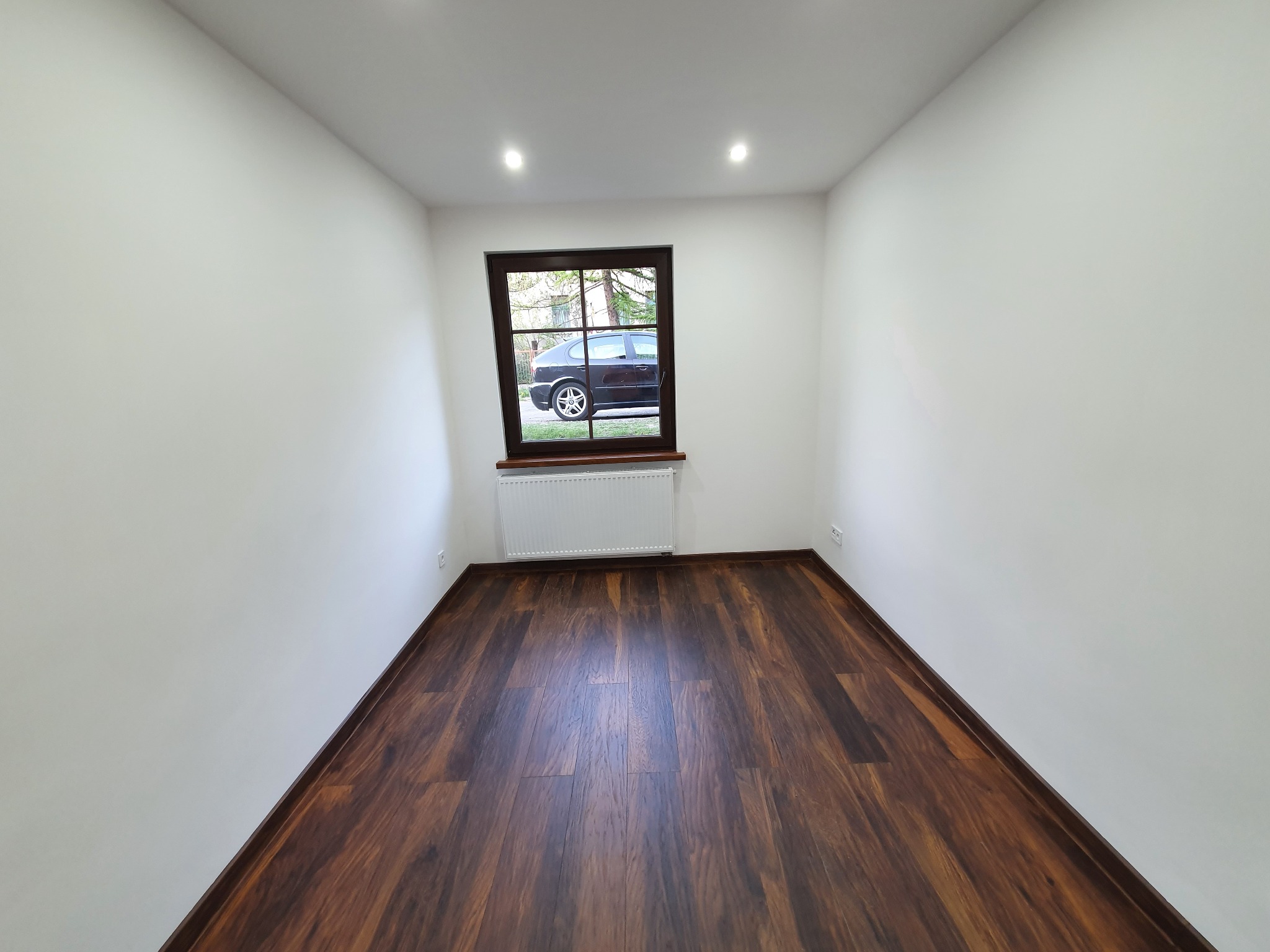 Pusty pokój po remoncie: świeżo pomalowane białe ściany, ciemna drewniana podłoga, okno z widokiem na samochód na zewnątrz, biały grzejnik pod oknem.