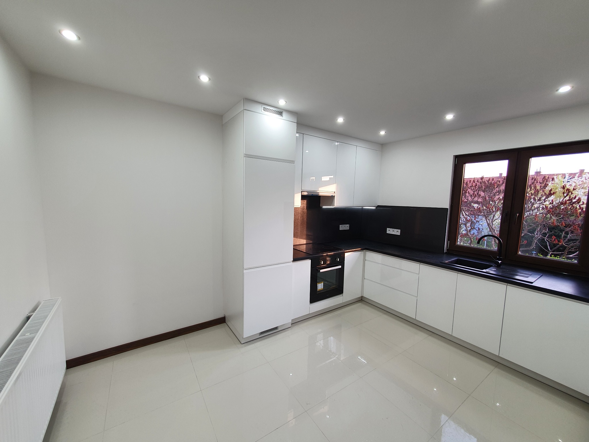 Nowoczesna, minimalistyczna kuchnia z białymi, lakierowanymi szafkami, czarnym blatem i widokiem na drzewo za oknem, oświetlona punktowymi lampami sufitowymi i z grzejnikiem żeberkowym w rogu.