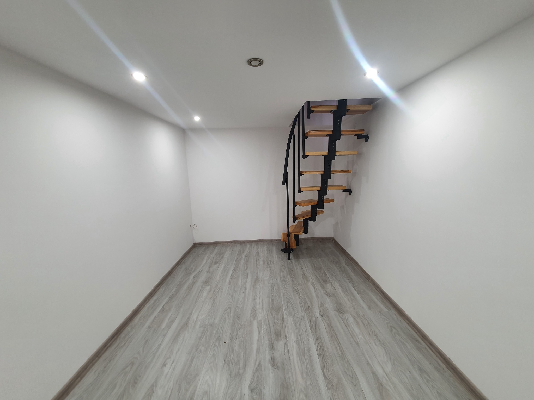 Puste, świeżo odnowione pomieszczenie z drewnianymi, spiralnymi schodami z czarną metalową konstrukcją, szarą podłogą imitującą drewno i białymi ścianami z widocznymi punktowymi lampami sufitowymi.