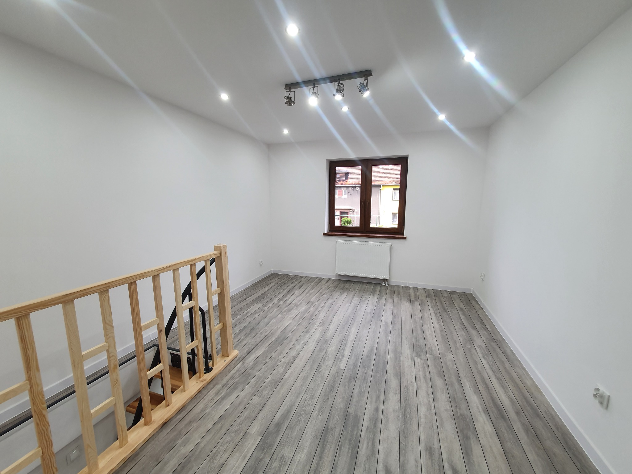 Jasne, puste pomieszczenie po remoncie z panelami podłogowymi imitującymi drewno, widoczne schody z drewnianą balustradą, białe ściany, okno z brązową ramą i kaloryfer pod nim, oraz oświetlenie...