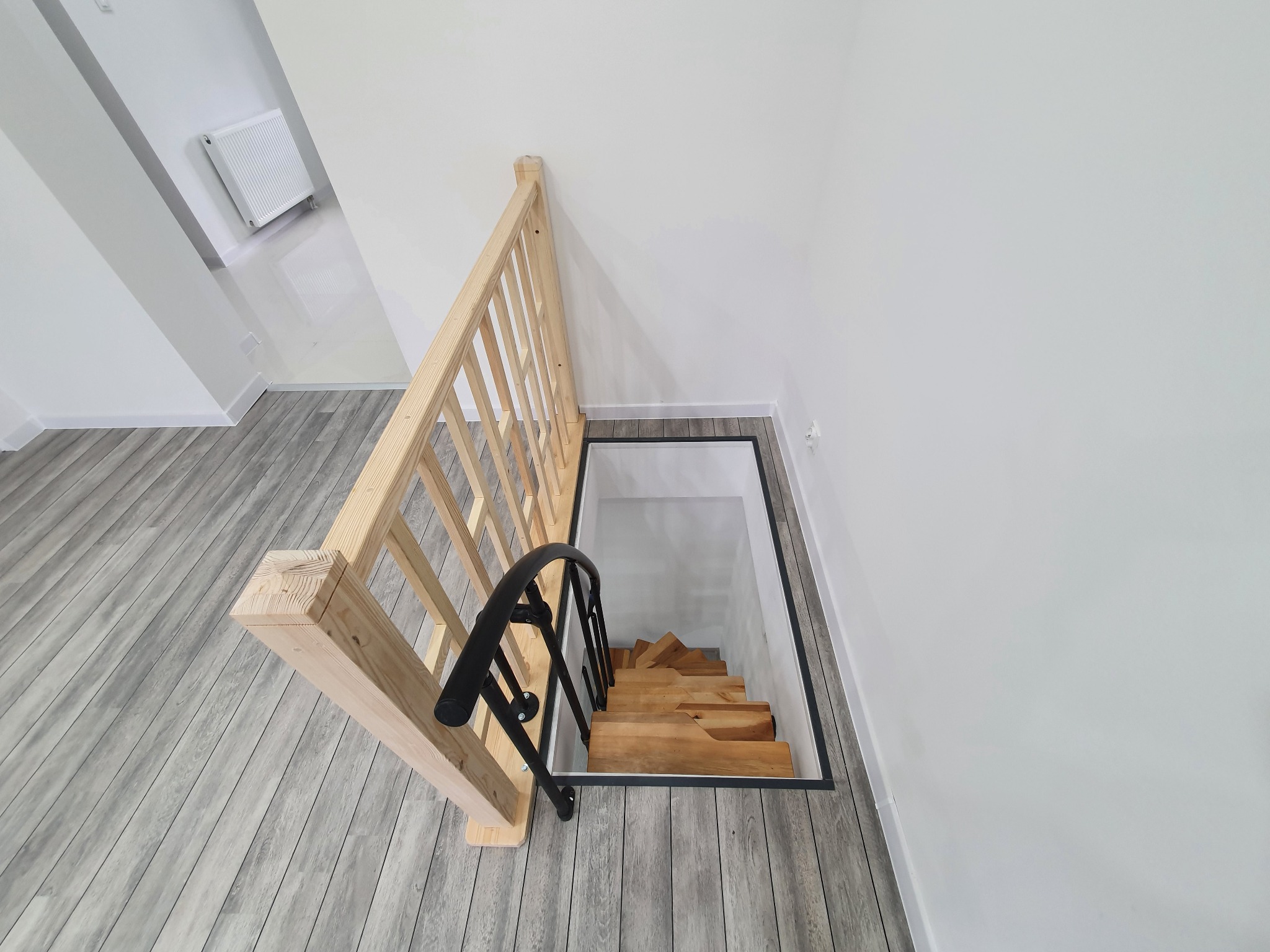 Widok z góry na drewniane schody prowadzące w dół, z jasną drewnianą balustradą i czarną metalową poręczą, na tle szarej podłogi imitującej drewno i białych ścian.