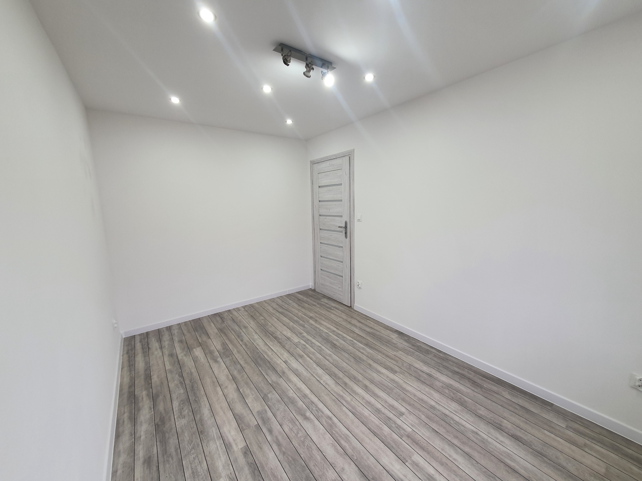 Pusty pokój po remoncie z szarymi panelami podłogowymi, białymi ścianami, drzwiami z poziomymi frezami i oświetleniem sufitowym halogenowym i reflektorowym.