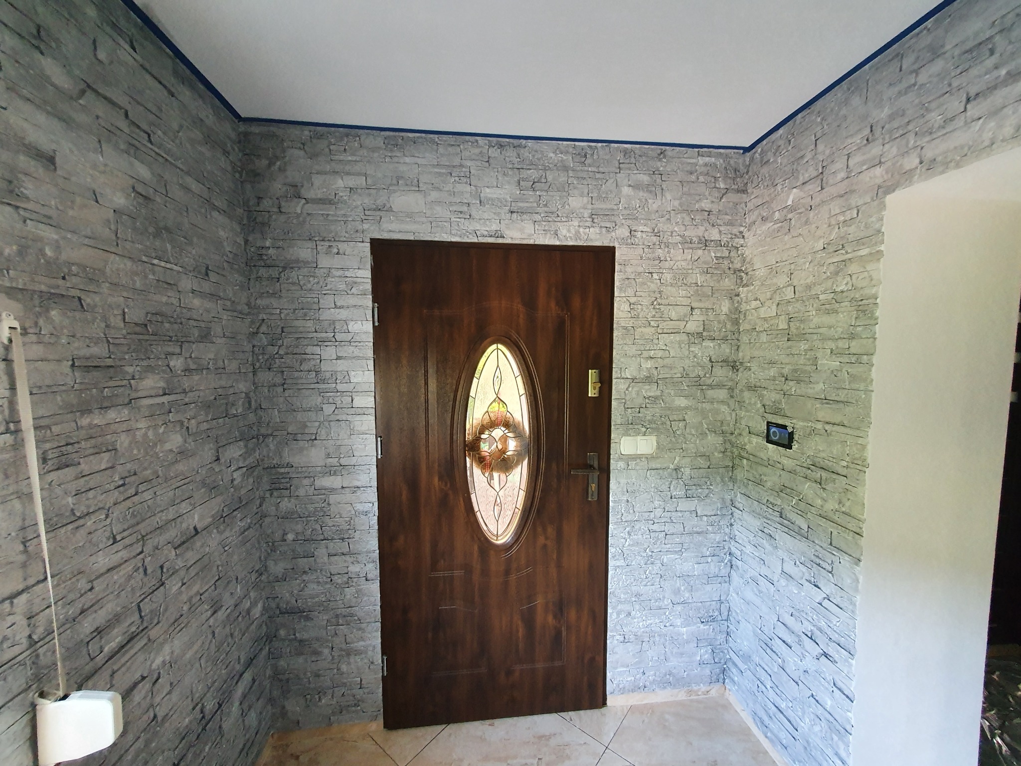 Przedpokój z dekoracyjną ścianą wykończoną szarą imitacją kamienia, drewniane drzwi wejściowe z owalnym przeszkleniem, widoczny panel sterowania na ścianie.