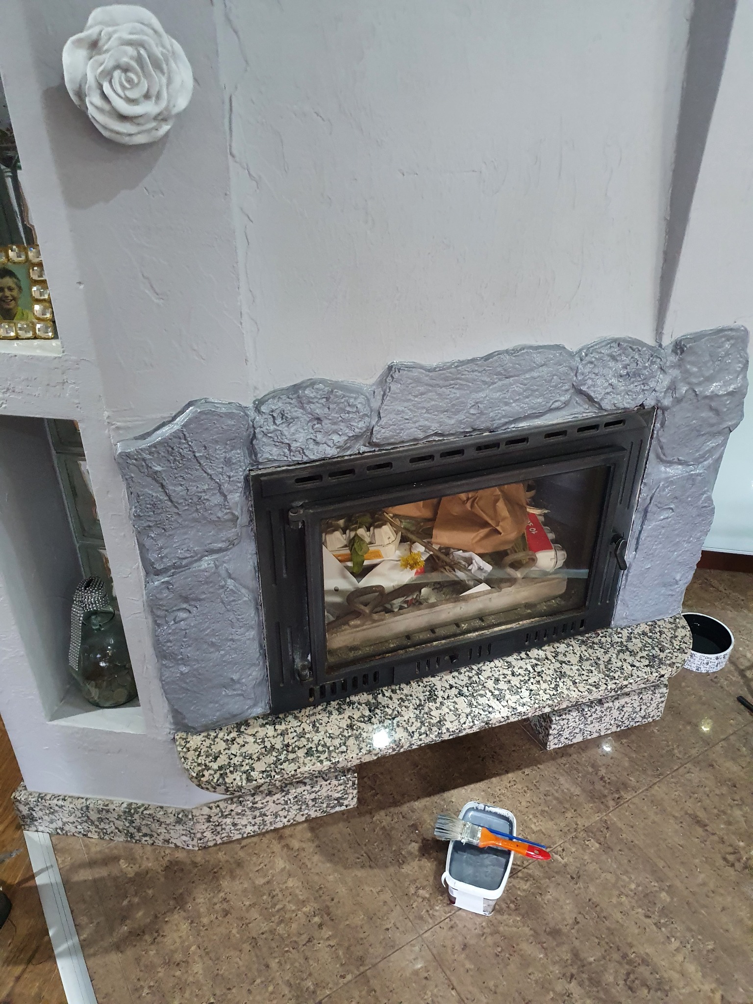 Otynkowany kominek z imitacją kamienia pomalowany na szaro, z farbą i pędzlem na podłodze, widoczny detal dekoracyjny w kształcie róży