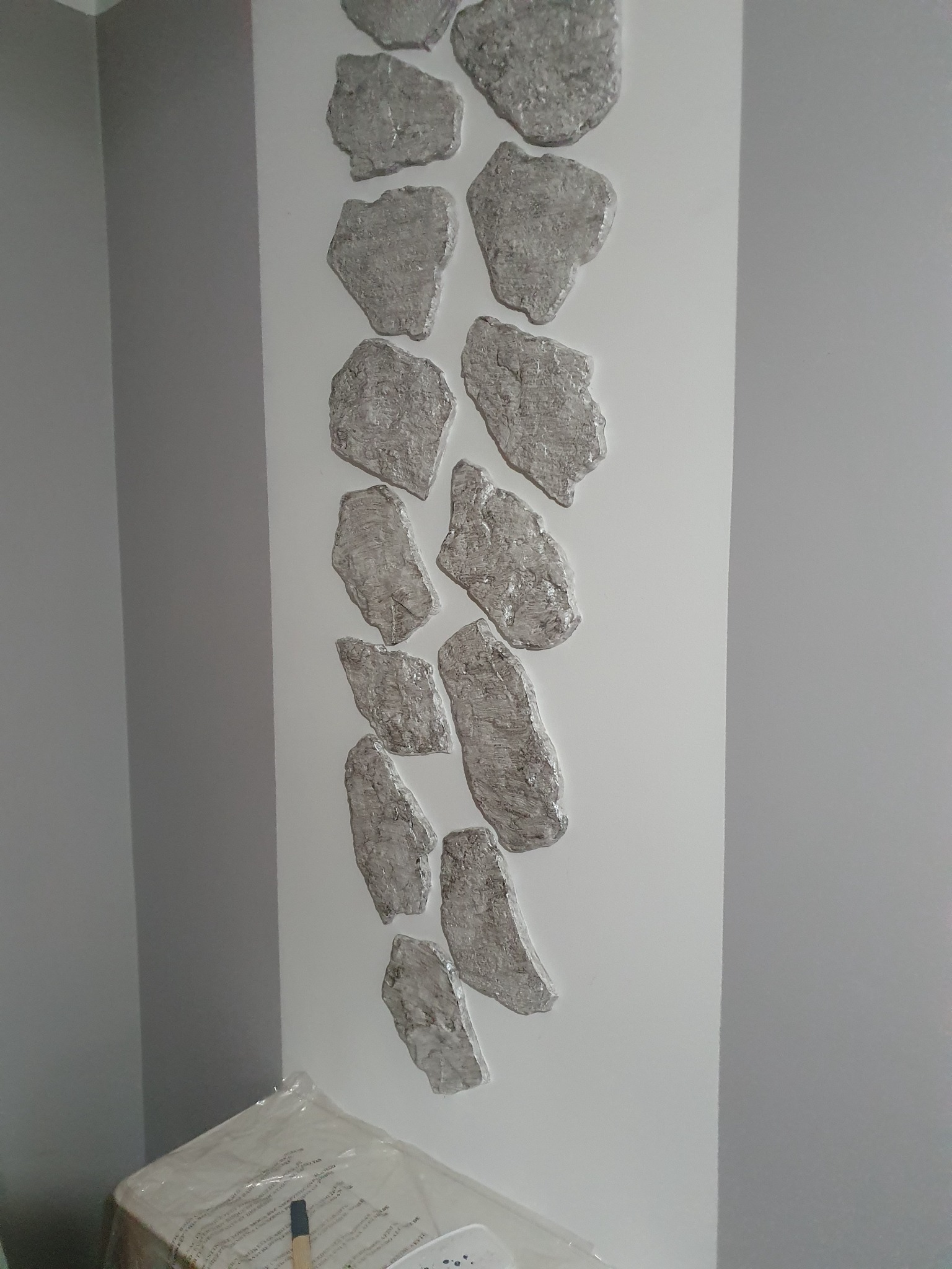 Dekoracyjna ściana z nałożoną strukturą przypominającą srebrne kamienie, ułożonych pionowo na białym tle, widoczny fragment narożnika pokoju pomalowanego na szaro.