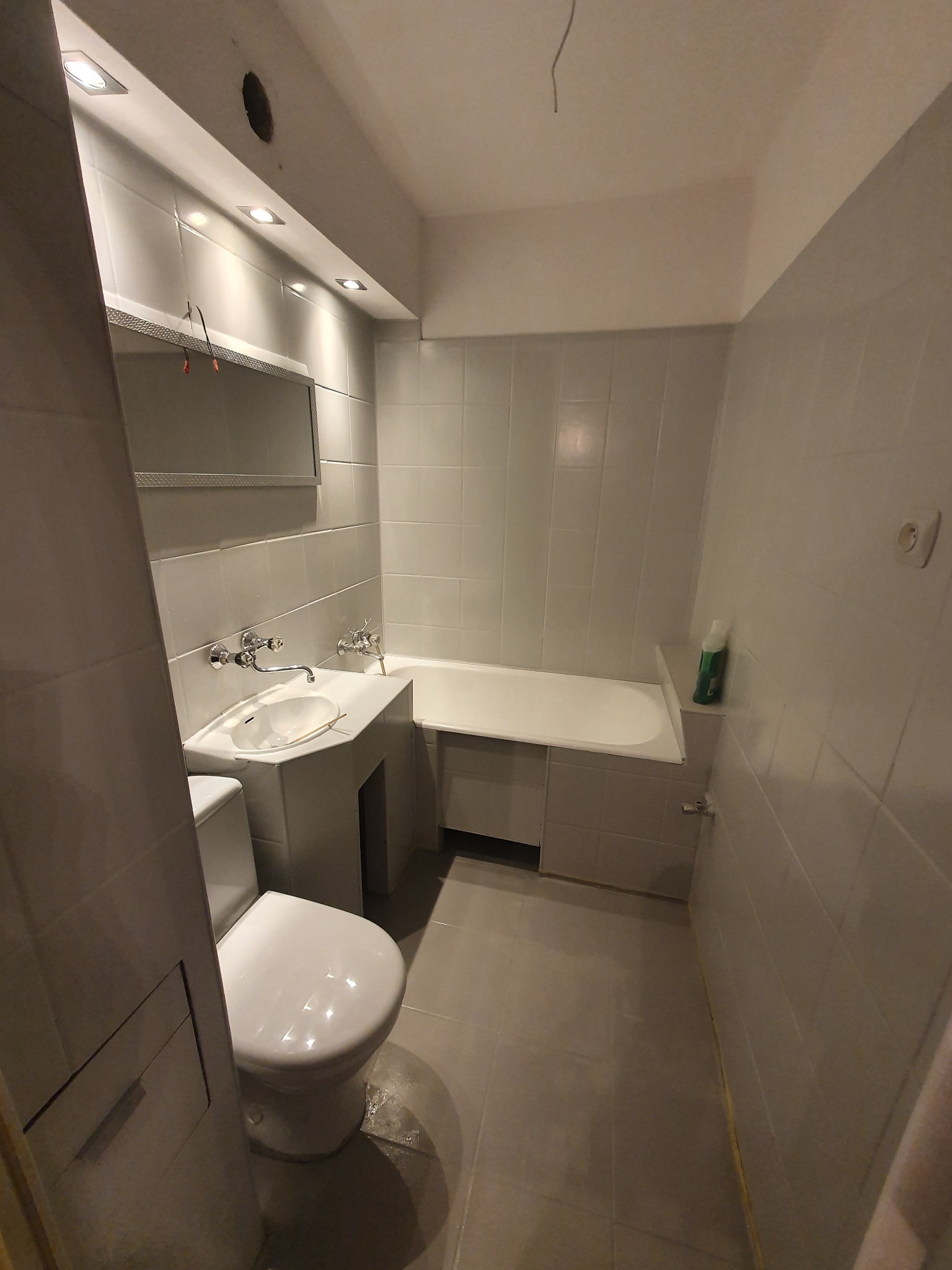 Szara łazienka z wanną, umywalką i toaletą, widoczne białe płytki na ścianach, sufit z oświetleniem punktowym i niedokończonym otworem, wnętrze w trakcie remontu.