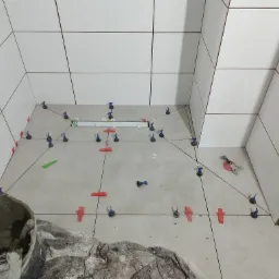 Układanie białych płytek w łazience z użyciem systemu poziomowania płytek, widoczny wiadro z zaprawą i narzędzia glazurnicze.