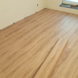 Fragment pokoju z ułożoną podłogą z paneli laminowanych o jasnym odcieniu drewna, widoczne nieużyte panele i niebieska taśma malarska przy oknie.