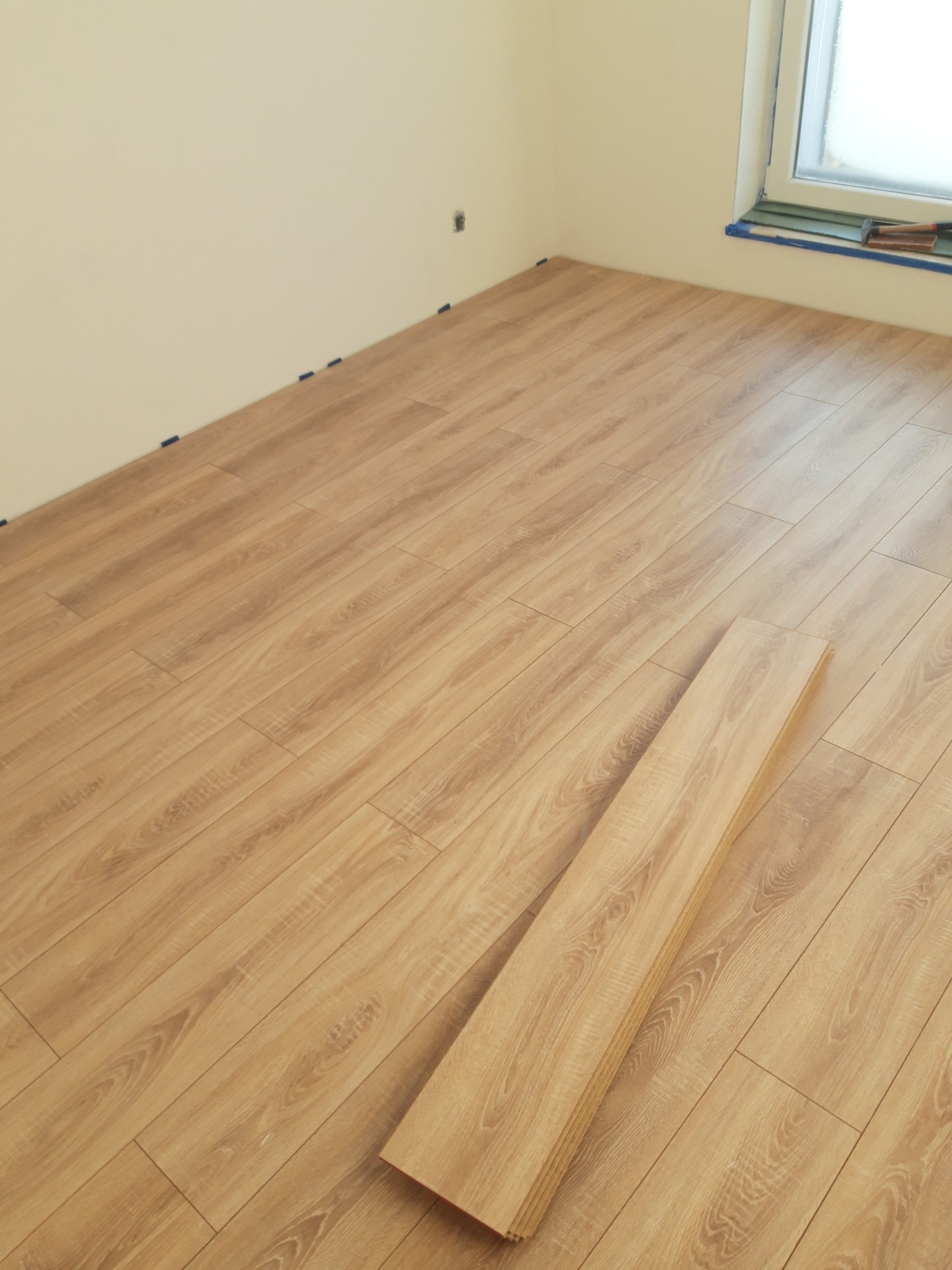 Fragment pokoju z ułożoną podłogą z paneli laminowanych o jasnym odcieniu drewna, widoczne nieużyte panele i niebieska taśma malarska przy oknie.