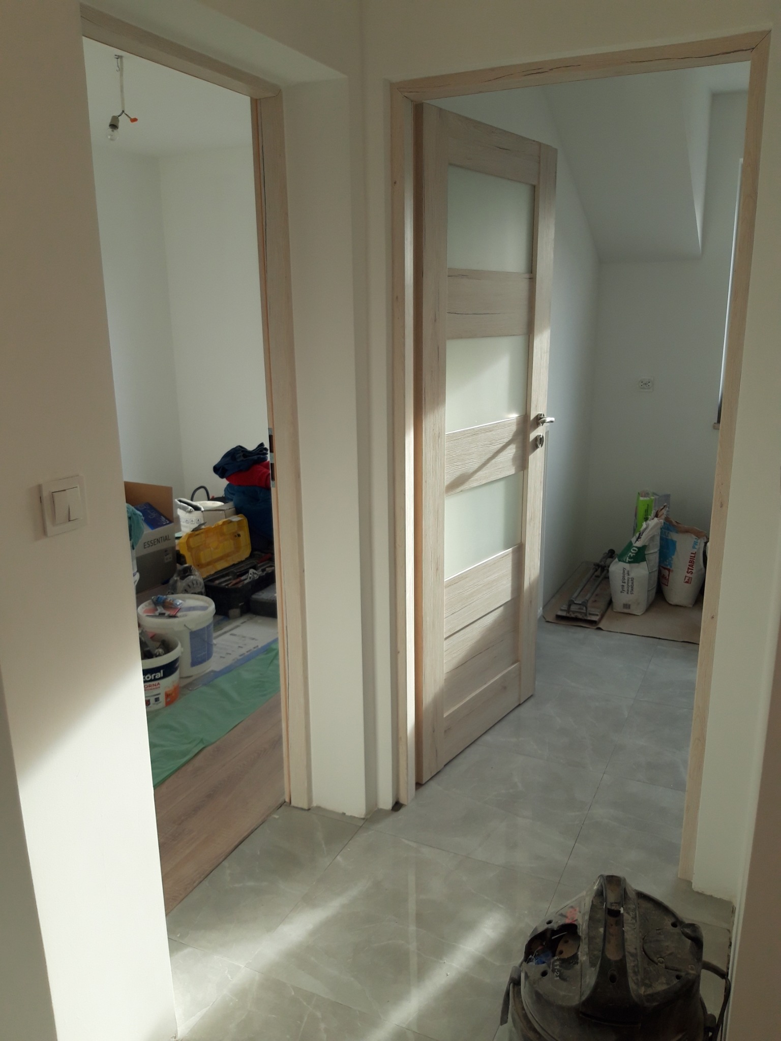 Dwa otwarte przejścia drzwiowe w trakcie prac wykończeniowych, widoczny fragment podłogi wyłożonej szarymi płytkami, w tle wiadro z farbą, torba z ubraniami i worki z zaprawą.