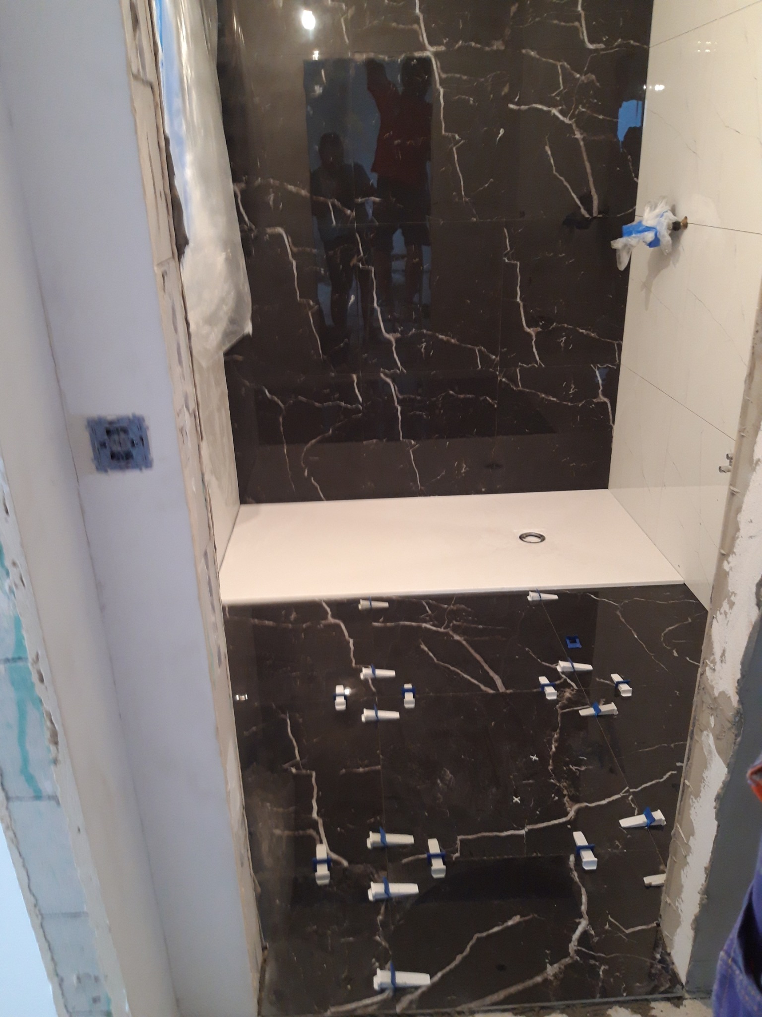 Łazienka w trakcie prac: Czarna marmurowa glazura na ścianach i podłodze, biały brodzik, plastikowe krzyżyki dystansowe między płytkami, widoczne niedokończone obramowania drzwi i instalacje.
