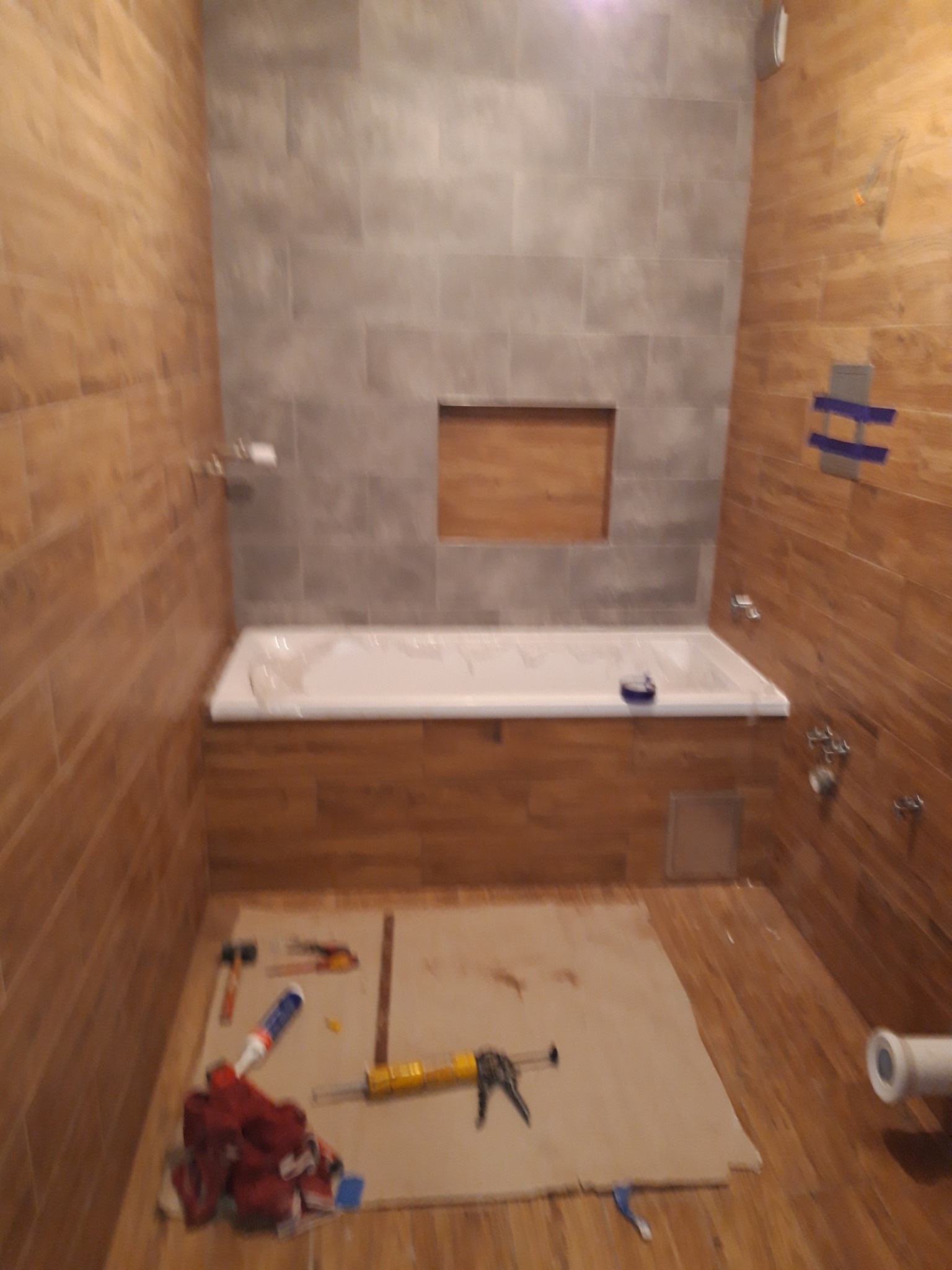 Wnętrze łazienki w trakcie remontu: widoczna wanna obudowana drewnopodobnymi płytkami, częściowo wyłożona szarymi płytkami ściana z wnęką, narzędzia i materiały budowlane na podłodze zabezpieczonej...