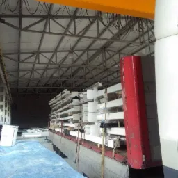 Części konstrukcji stalowej, pomalowane na biało, zabezpieczone folią i załadowane na ciężarówkę wewnątrz hali magazynowej, widoczne elementy suwnicy pod stropem.