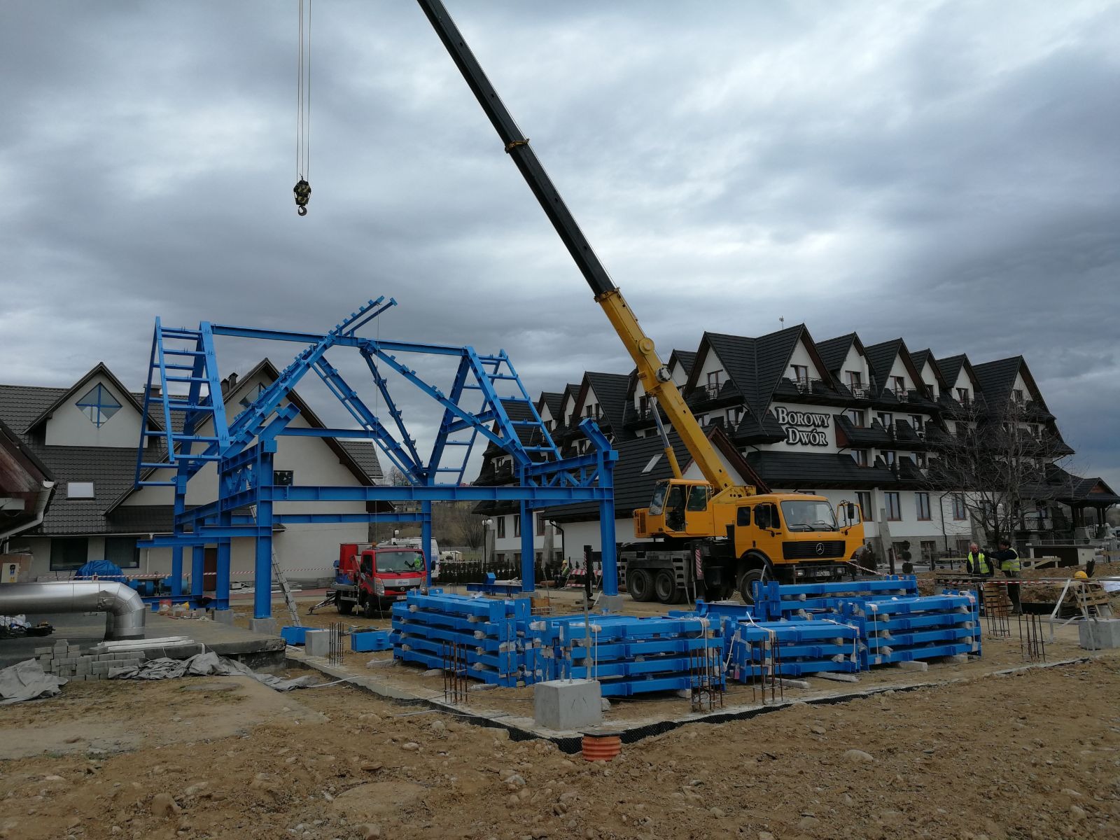 Montaż niebieskiej konstrukcji stalowej hali z użyciem żółtego dźwigu na tle budynku Borowy Dwór.