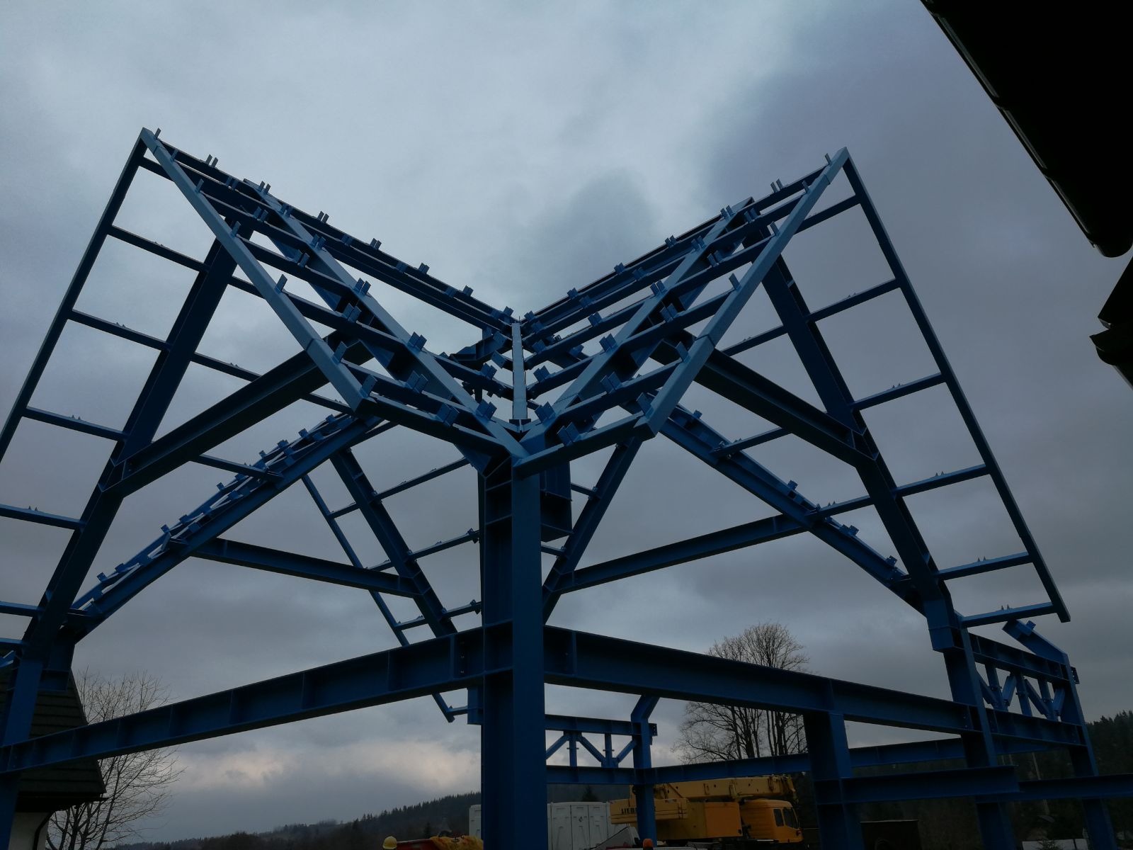 Niebieska stalowa konstrukcja hali w trakcie montażu na tle pochmurnego nieba, widoczny żuraw budowlany w tle.