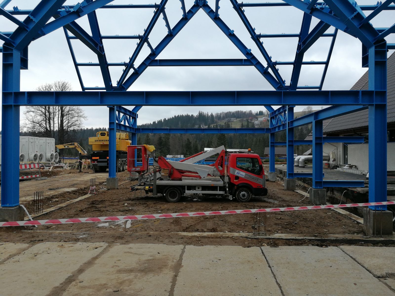 Montaż niebieskiej konstrukcji stalowej hali w Niedomicach, widoczny dźwig samochodowy i teren budowy z wykopami.