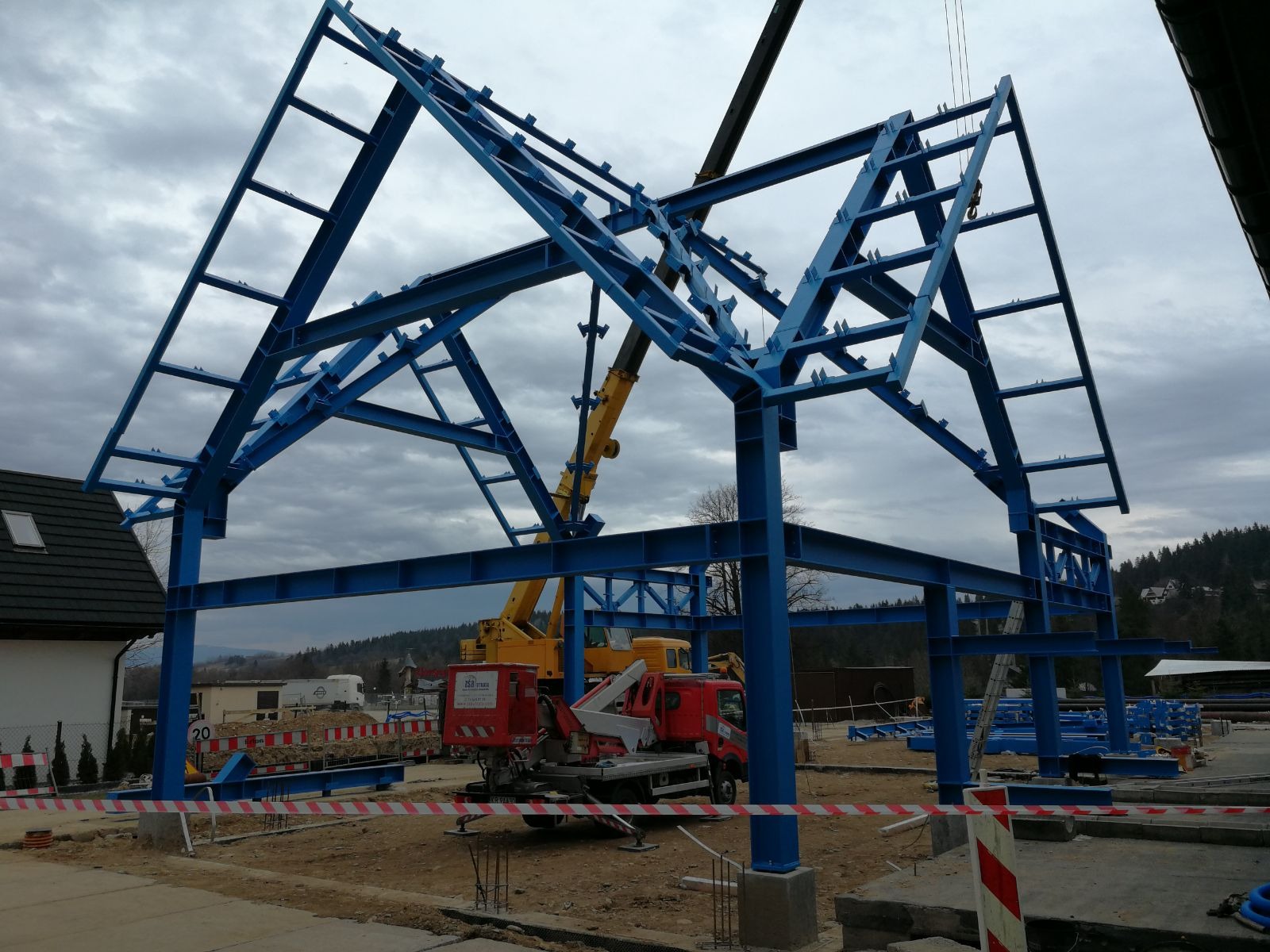 Stalowa konstrukcja szkieletowa budynku w kolorze niebieskim, w trakcie montażu przez dźwig na placu budowy z widocznymi elementami zabezpieczeń i sprzętem budowlanym.