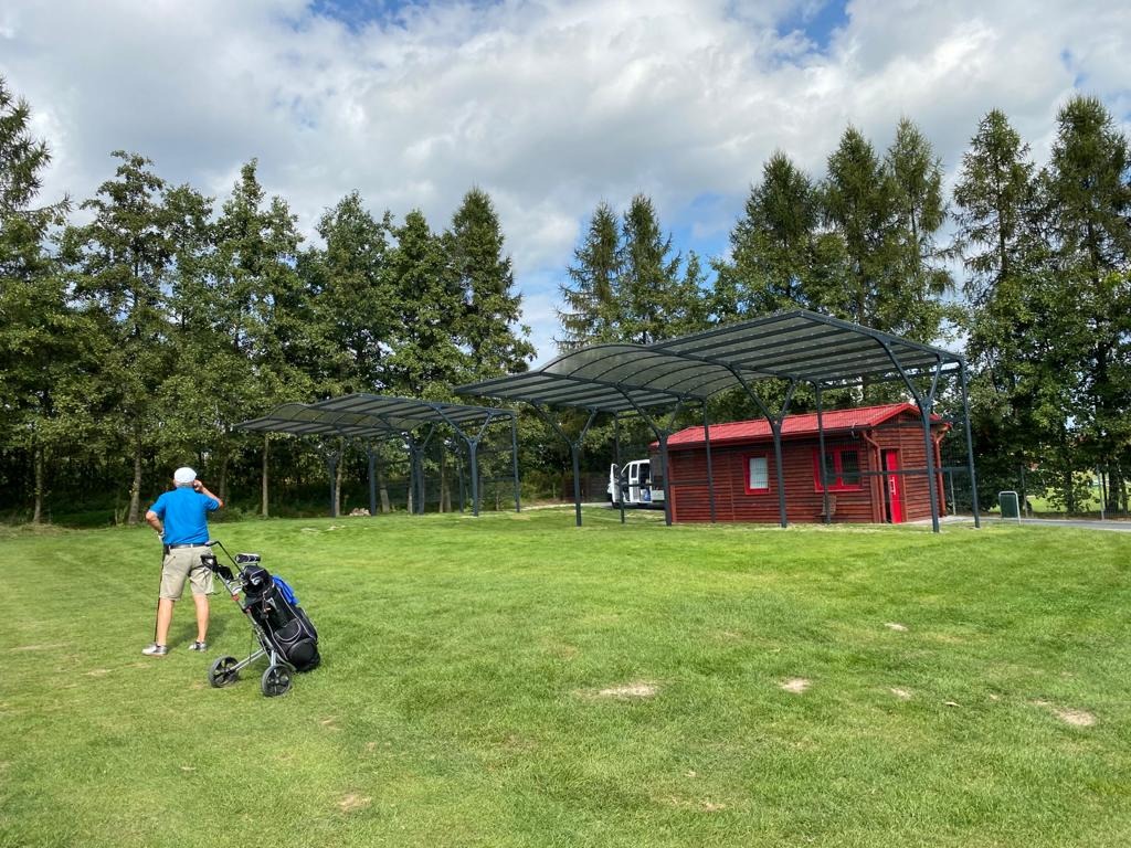 Zadaszenia z metalową konstrukcją i przezroczystym dachem nad polem golfowym, obok czerwony budynek gospodarczy z białymi oknami i czerwonymi drzwiami, w tle wysokie drzewa i błękitne niebo...