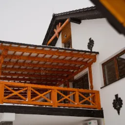 Nowoczesny dom z drewnianym balkonem i dekoracyjnym elementem metalowym na białej elewacji podczas opadów śniegu.