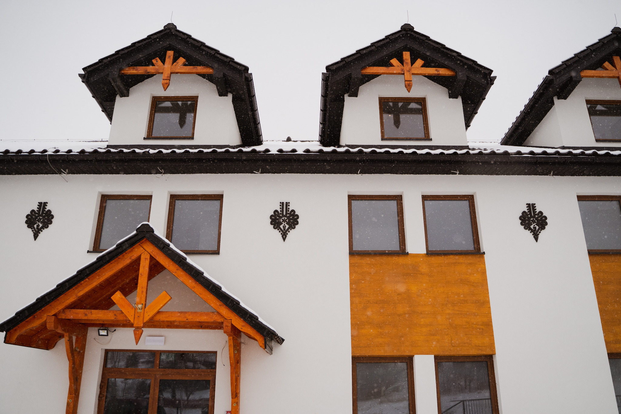 Fasada budynku z białymi ścianami, drewnianymi oknami i elementami dekoracyjnymi w stylu góralskim, pokryta śniegiem.