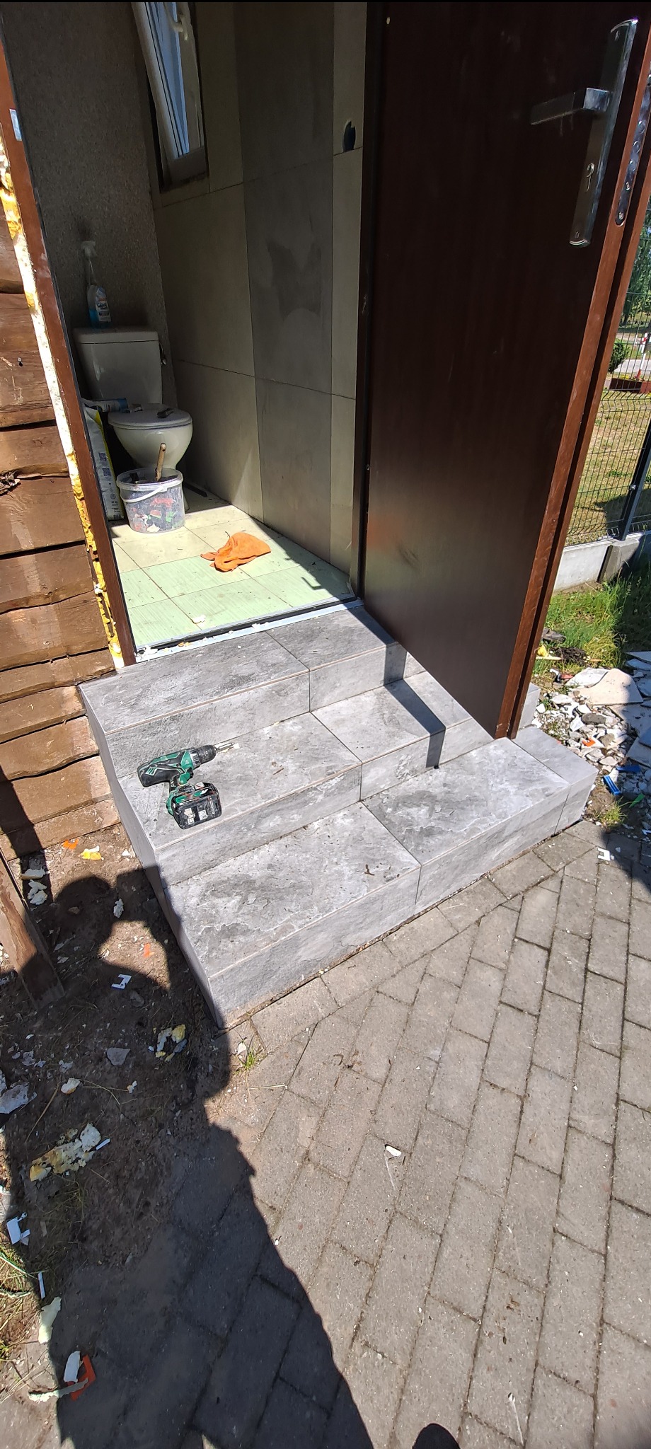 Wejście do toalety w domku letniskowym z trzema schodkami wyłożonymi szarymi płytkami; na schodach leży wkrętarka akumulatorowa; wnętrze toalety wyłożone jasnymi płytkami.