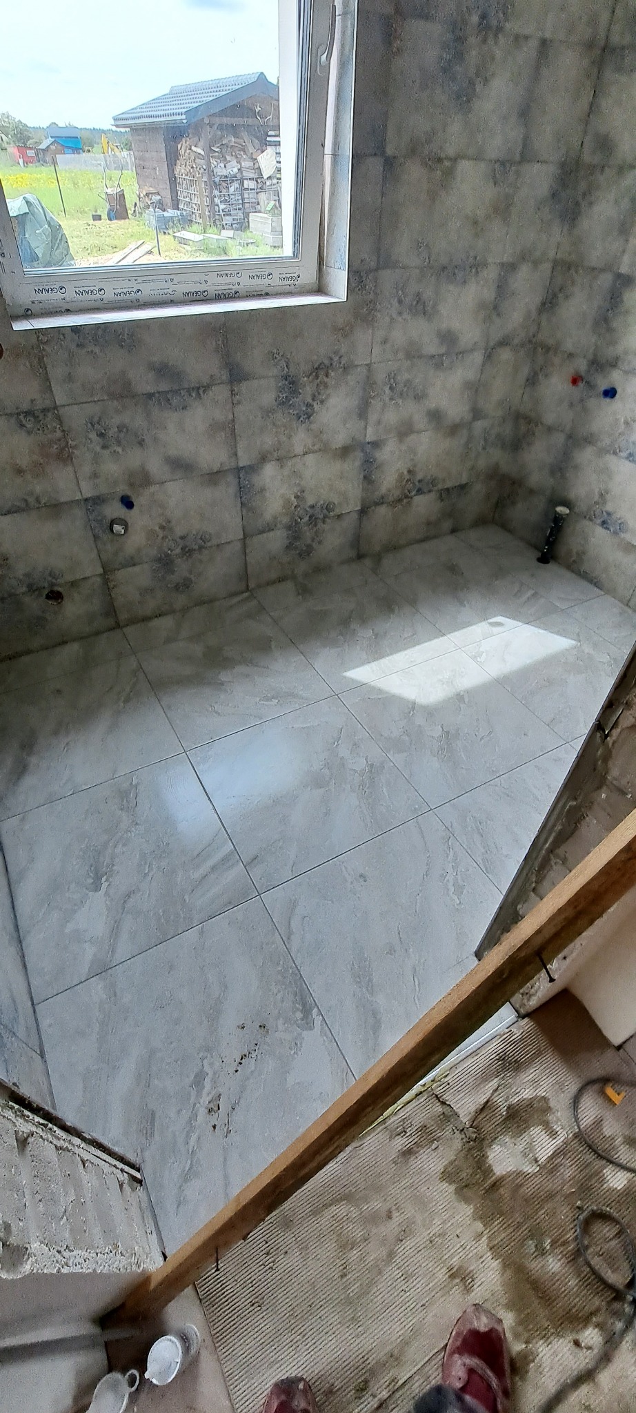 Wykończona łazienka z szarymi płytkami imitującymi kamień, widoczne przygotowania do dalszych prac wykończeniowych: listwa przypodłogowa, narzędzia, widok przez okno na podwórko.
