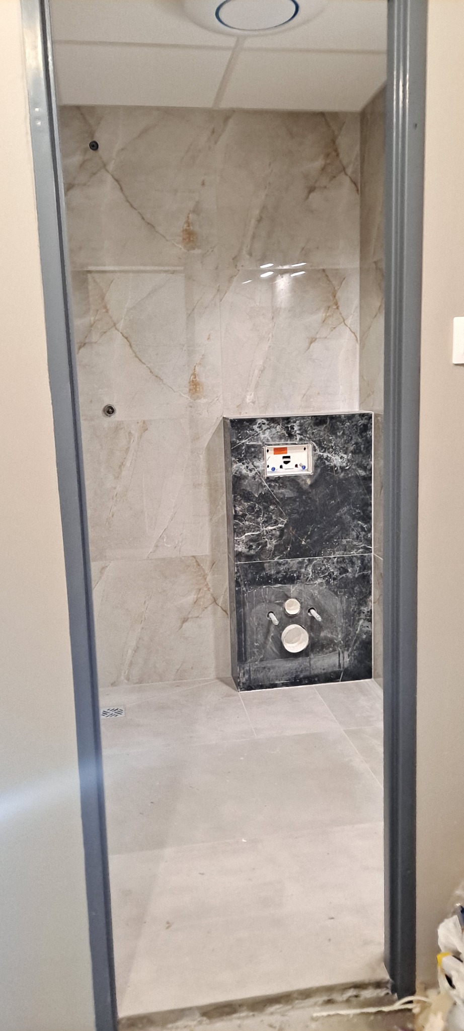 Wykończona łazienka z beżowymi marmurowymi płytkami na ścianach i czarnym panelem maskującym instalację sanitarną, widok przez futrynę drzwi.