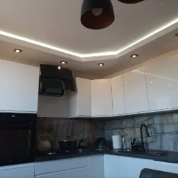 Nowoczesna kuchnia z białymi, błyszczącymi szafkami, czarnym okapem i piekarnikiem, blatem w kolorze antracytu oraz dekoracyjnym oświetleniem LED podszafkowym i sufitowym.