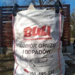 Worek Big Bag z napisem 'Odbiór gruzu i odpadów' stojący na trawie przed brązowym ogrodzeniem i drzewami.