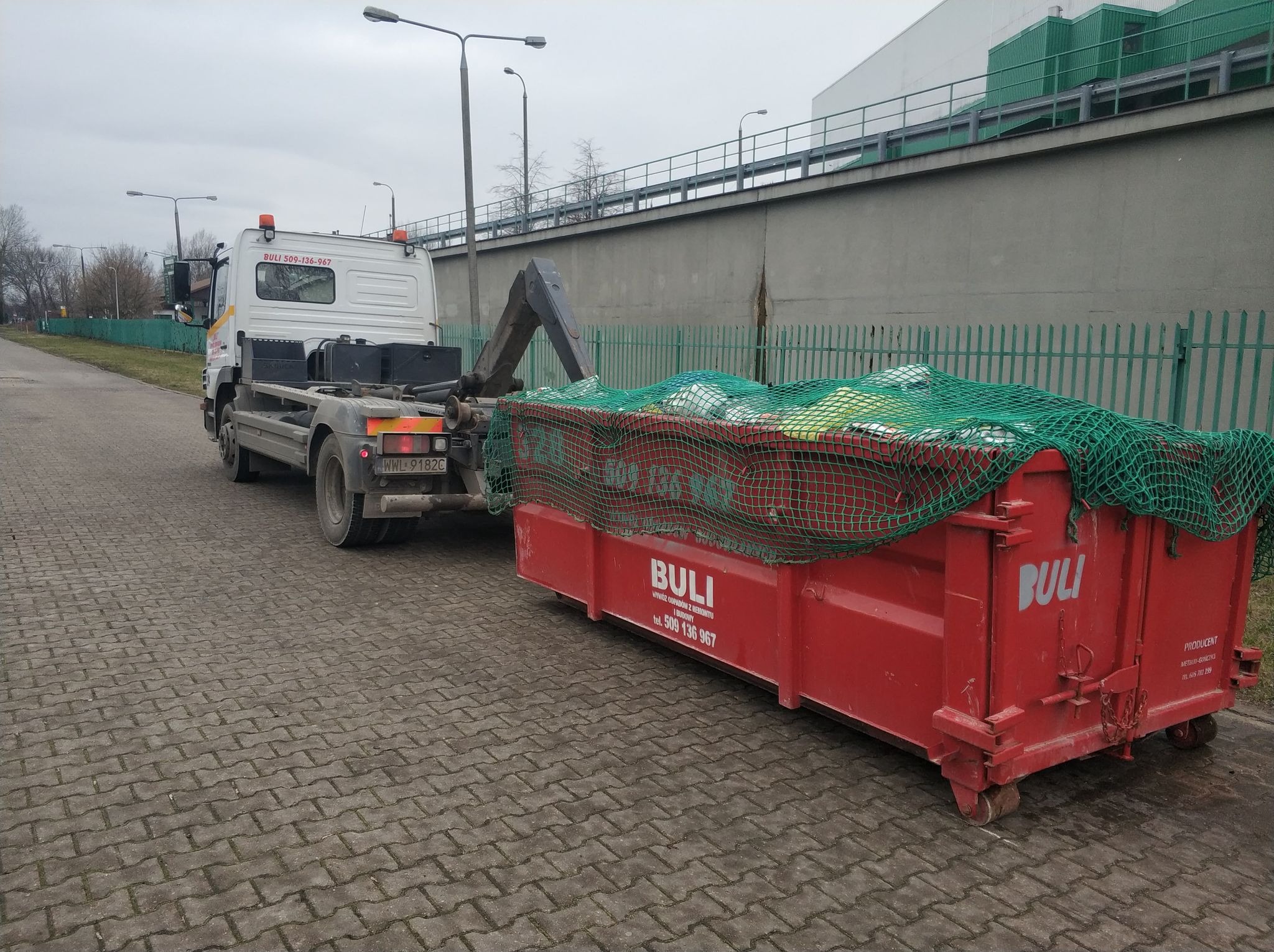 Czerwony kontener na gruz marki Buli, zabezpieczony zieloną siatką, załadowany na ciężarówkę z dźwigiem na kostce brukowej, w tle szary budynek przemysłowy i zielone ogrodzenie.