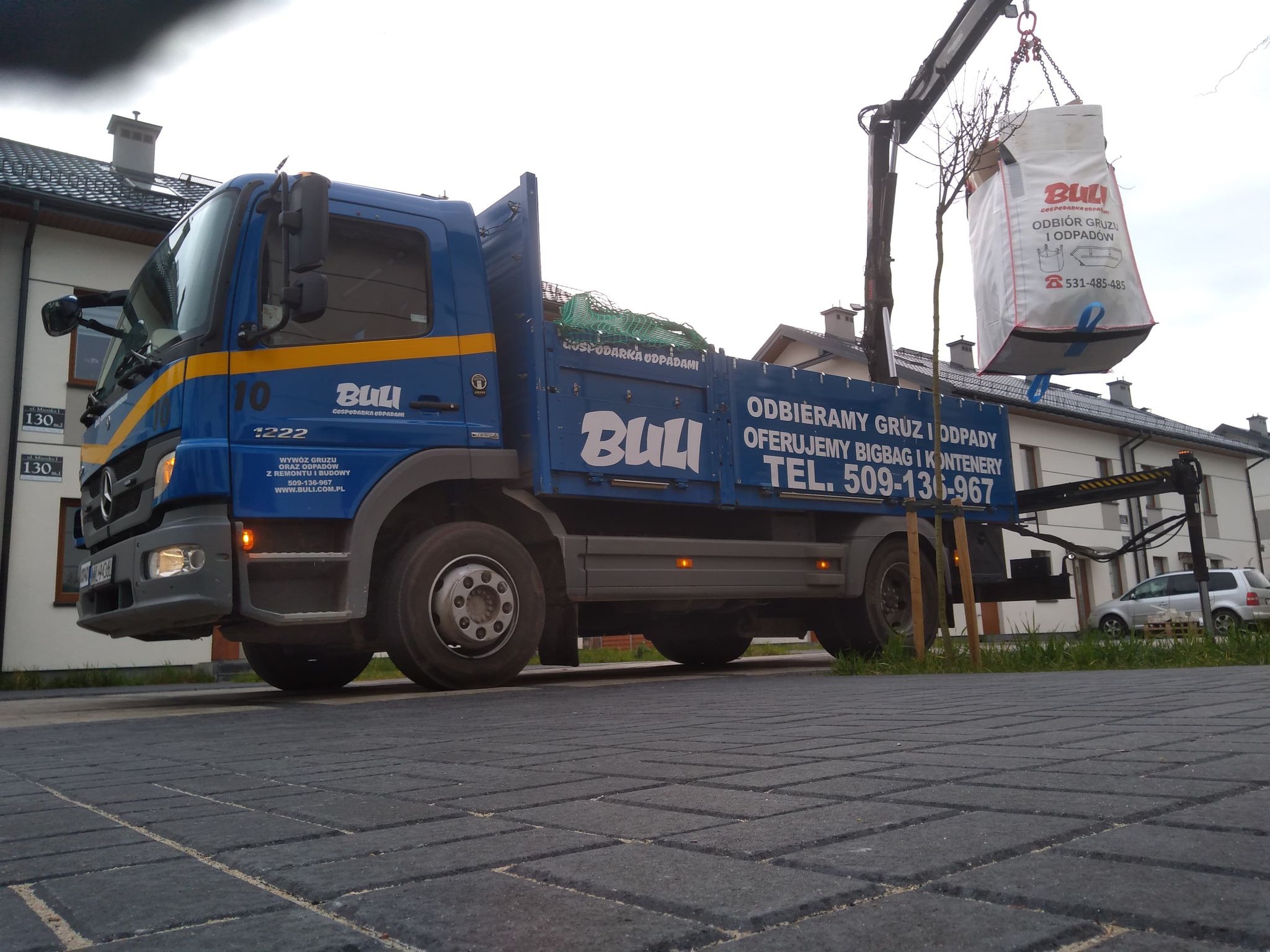 Niebieska ciężarówka z dźwigiem podnosi duży worek z napisem 'Odbiór gruzu i odpadów' na tle budynków mieszkalnych.