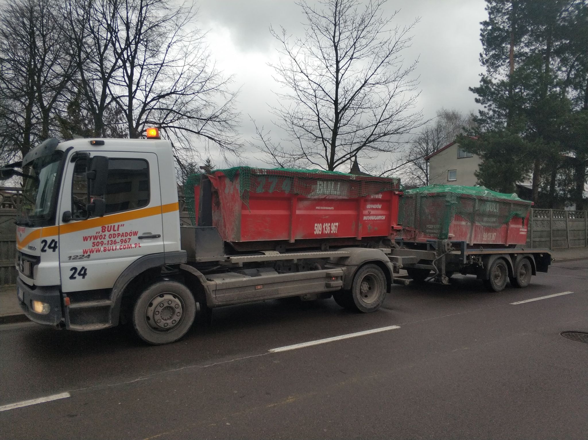 Ciężarówka z przyczepą przewożąca dwa czerwone, częściowo zakryte siatką, kontenery. Na kabinie ciężarówki widoczne logo firmy zajmującej się wywozem odpadów. Tło stanowią drzewa i budynki mieszkalne.