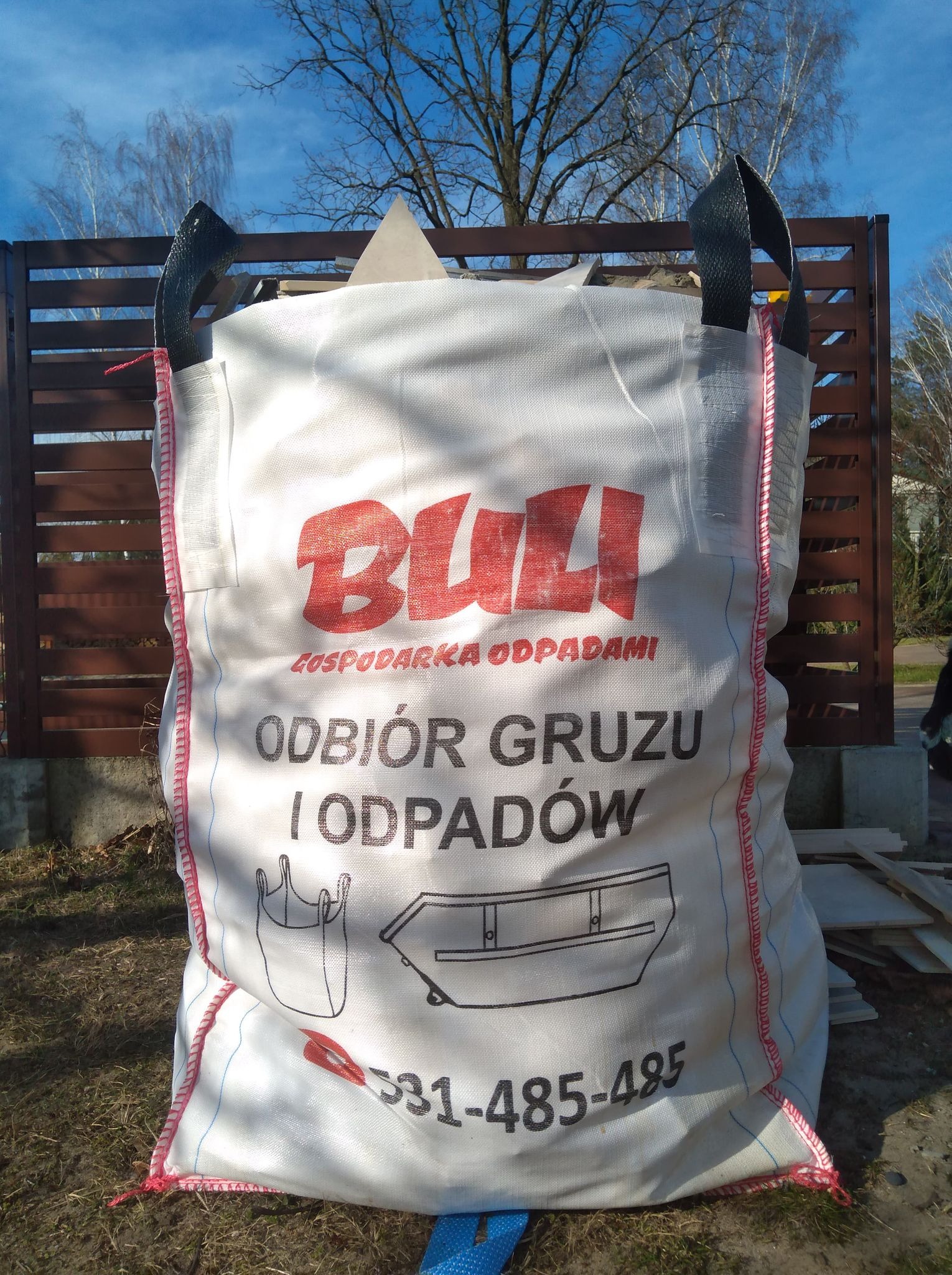 Worek Big Bag z napisem 'Odbiór gruzu i odpadów' stojący na trawie przed brązowym ogrodzeniem i drzewami.