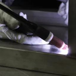Zbliżenie na proces łączenia metalowych elementów za pomocą metody TIG, widoczny jasny łuk elektryczny i spawany narożnik profilu, rękawica ochronna w tle.