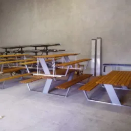 Metalowe konstrukcje stołów piknikowych z drewnianymi blatami i siedziskami w szarej hali produkcyjnej, z widocznymi elementami metalowymi w tle.