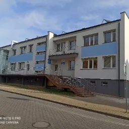 Dwupiętrowy budynek z jasną elewacją, niebieskimi akcentami i zewnętrzną windą, widoczne okna z żaluzjami, schody z metalową balustradą prowadzące do wejścia.