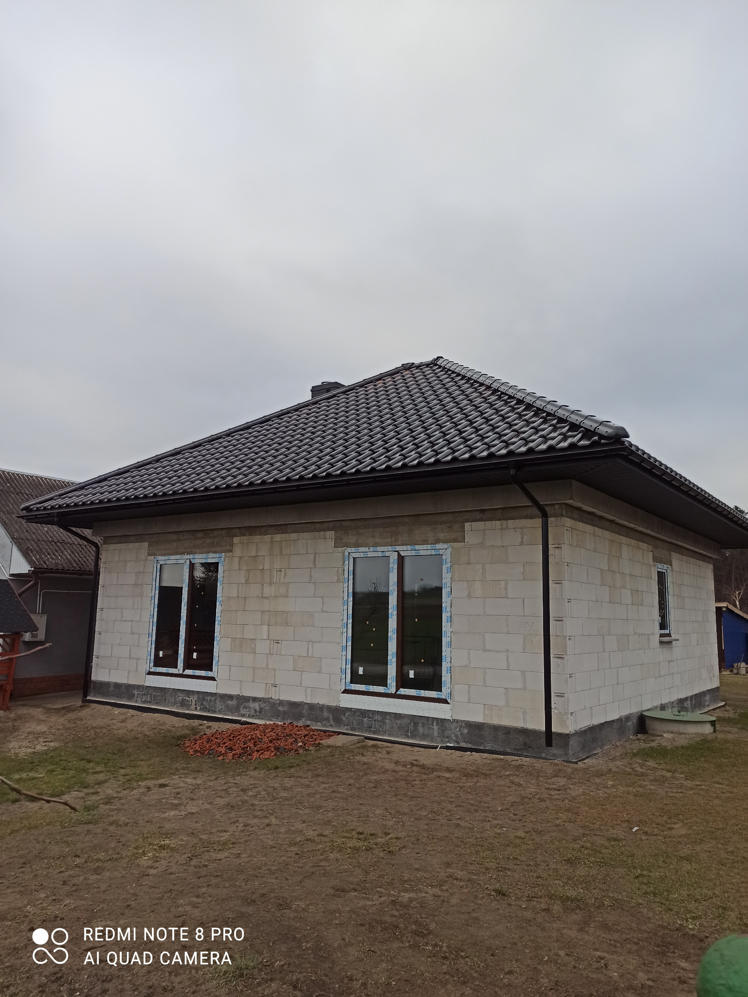 Surowy stan budynku z białego bloczka z zamontowanymi oknami z niebieską folią ochronną, czarny dach z dachówki ceramicznej, widoczne pozostałości po pracach budowlanych w postaci sterty cegieł...