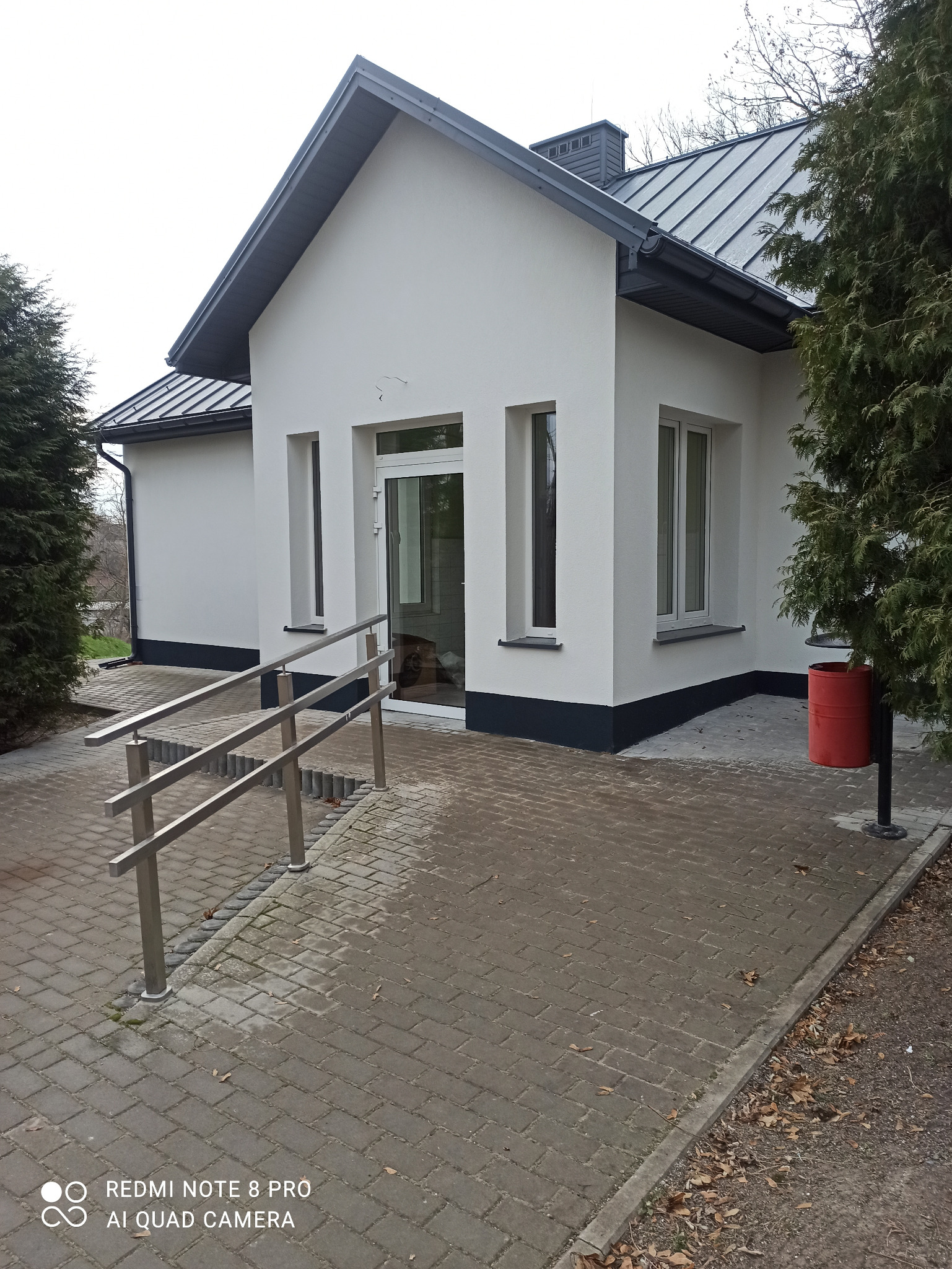 Nowoczesny budynek z białymi ścianami, szarym dachem i srebrnymi poręczami przy wejściu, widok z zewnątrz, pochmurny dzień.