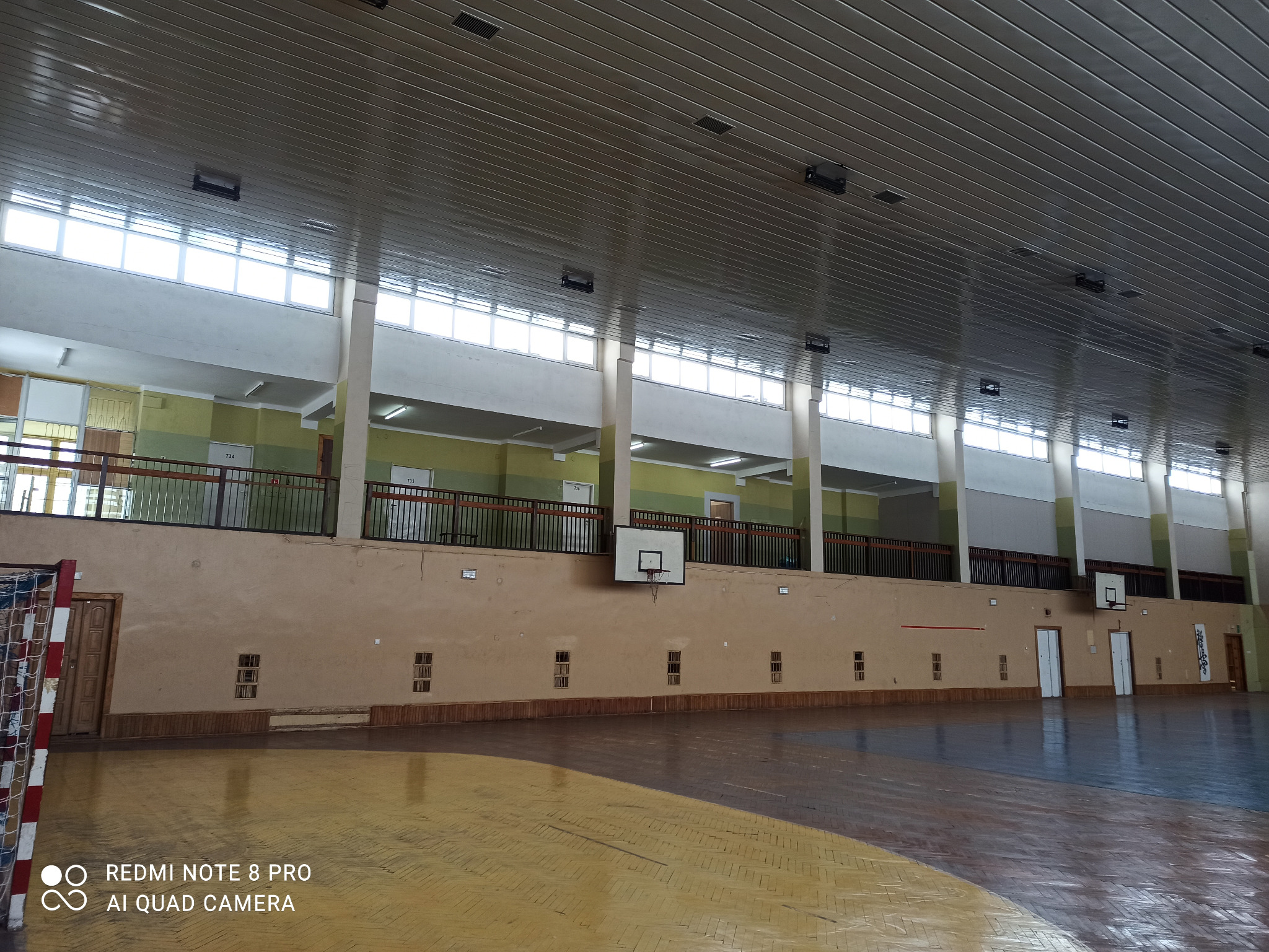Wnętrze hali sportowej z parkietem, widoczne okna PCV pod sufitem, oświetlenie, kosze do koszykówki na ścianie i siatka do piłki ręcznej.