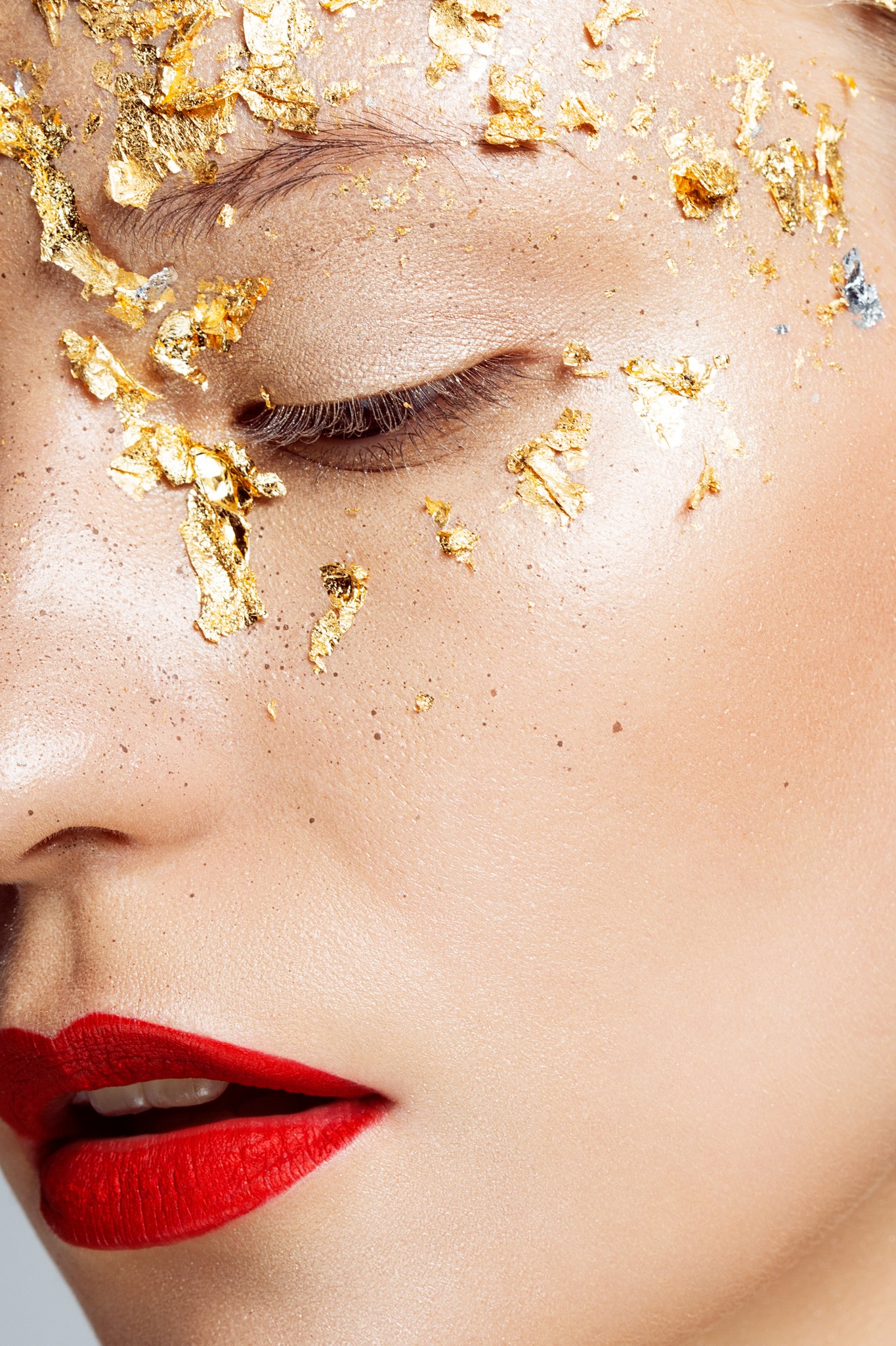 Zbliżenie na twarz kobiety z zamkniętym okiem, pokrytą złotymi płatkami i intensywnie czerwoną szminką na ustach.