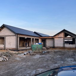 RASBUD SPÓŁKA Z OGRANICZONĄ ODPOWIEDZIALNOŚCIĄ - Budowa domu jednorodzinnego w stanie surowym otwartym: ściany z białego bloczka, dach pokryty ciemną dachówką, widoczny kontener na gruz i materiały budowlane.