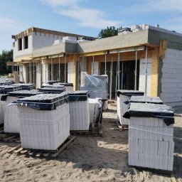 RASBUD SPÓŁKA Z OGRANICZONĄ ODPOWIEDZIALNOŚCIĄ - Budowa domu jednorodzinnego w Dziwnowie. Widoczne ściany z białych bloczków, podpory stropu i palety z materiałami budowlanymi. Częściowo zrealizowany etap murowania ścian.