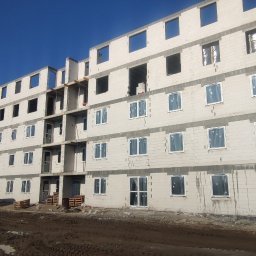 RASBUD SPÓŁKA Z OGRANICZONĄ ODPOWIEDZIALNOŚCIĄ - Wielopiętrowy budynek w trakcie budowy, z widocznymi oknami w folii ochronnej i białymi ścianami z betonu komórkowego, na tle błękitnego nieba i krajobrazu.